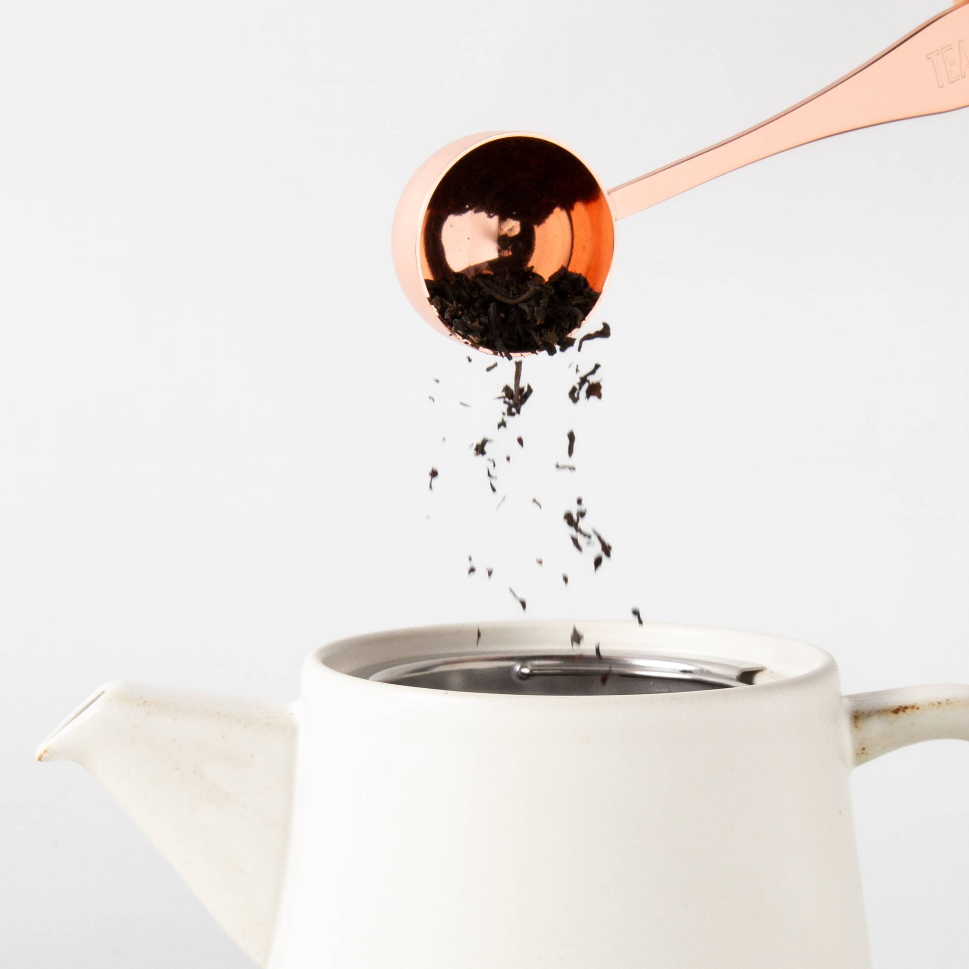 Copper tea scoop measuring loose leaf tea over a white ceramic teapot with strainer